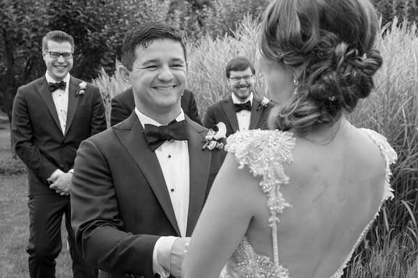 A black-and-white portrait-style shot capturing what appears to be a first look or outdoor ceremony moment, with the groom facing the camera wearing a dark tuxedo with bow tie and a small boutonniere, smiling broadly as he holds the bride's hands. The bride is seen from behind, her low-cut back dress featuring intricate lace or beaded detail, with her hair styled in an updo with small accessories. Two groomsmen in matching dark tuxedos and bow ties stand smiling in the soft-focus background. The setting appears to be an outdoor garden or grounds area with tall ornamental grasses and foliage visible. The image has strong mid-tones with good contrast, shot at a slight angle as a candid portrait.