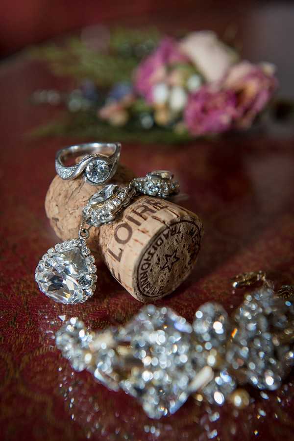 A close-up detail shot of bridal jewelry arranged around a Loire Valley wine cork. A solitaire diamond engagement ring and a diamond-set wedding band are balanced on top of the cork, while a large cushion-cut crystal drop earring with a halo of smaller stones leans against its side. A crystal-encrusted bracelet or necklace is blurred in the foreground, and a bouquet of pink, cream, and mauve flowers is softly out of focus in the background. The items are arranged on a deep red fabric surface, and the overall warm, dim lighting gives the image a rich, moody tone.