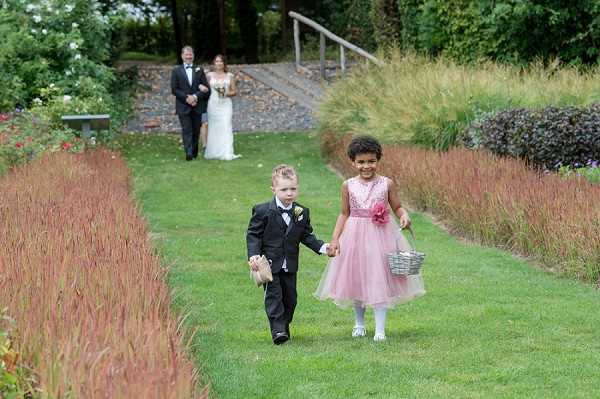 An outdoor garden ceremony procession showing a young ring bearer in a black tuxedo with a white boutonniere and a flower girl in a blush pink tulle dress with a pink flower sash, holding hands and walking down a grass aisle toward the camera; the flower girl carries a small silver wire basket. In the background, a groom in a black tuxedo and a bride in a white fitted gown holding a bouquet stand at the top of a stone staircase, waiting to begin their processional. The garden setting features ornamental grasses and red-toned border plantings along the aisle path. Wide shot capturing the full scene from the children in the foreground to the couple in the background.