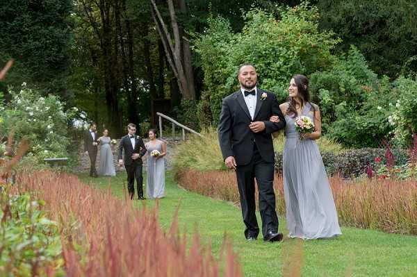 The bridal party is processing along a garden path during what appears to be a wedding ceremony procession. The setting is an outdoor formal garden with ornamental grasses, red-toned foliage borders, and mature trees in the background. In the foreground, a groomsman in a black tuxedo with bow tie and boutonniere walks arm-in-arm with a bridesmaid in a floor-length light grey chiffon dress carrying a small mixed bouquet with coral and yellow blooms. Behind them, two more groomsmen in black tuxedos escort two additional bridesmaids in matching light grey dresses, with another couple visible further back. The shot is a medium wide angle taken from ground level along the garden path.