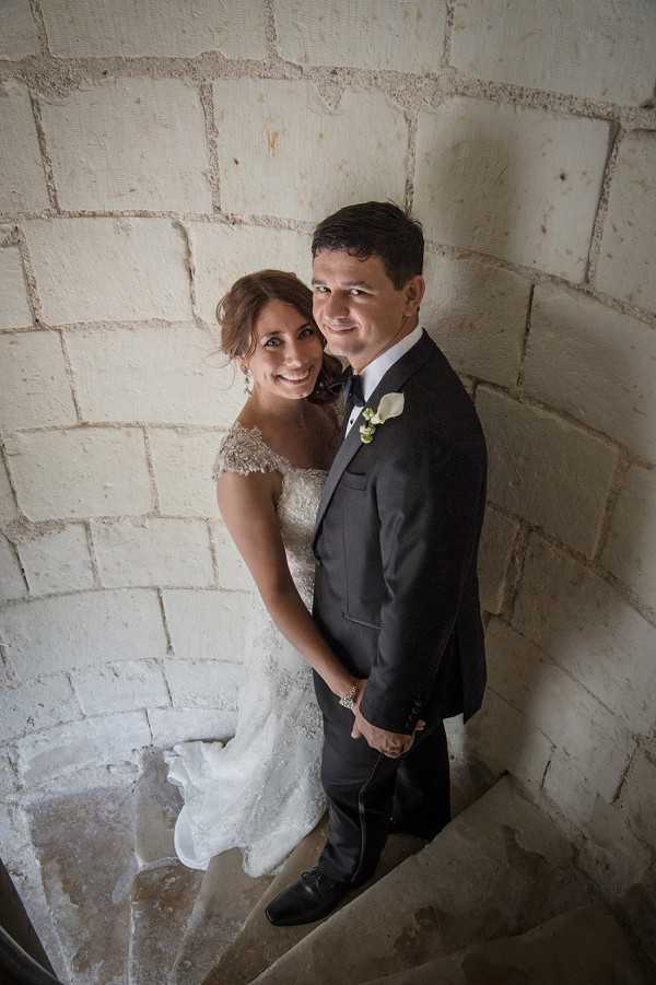 A couple portrait taken from an elevated angle looking down at a bride and groom standing together on a narrow stone spiral staircase inside what appears to be a chateau or historic tower. The bride wears an ivory lace gown with cap sleeves and has her hair styled in an updo, while the groom wears a charcoal suit with a white dress shirt, black tie, and a white calla lily boutonniere. The couple is embracing and smiling up toward the camera. The surrounding walls are composed of large, pale limestone blocks, giving the setting a classic French château character. The overhead perspective creates a slightly wide-angle portrait composition.