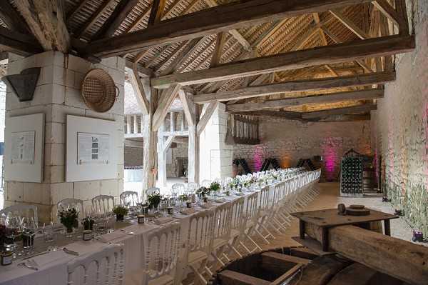 A wide-angle interior shot of a rustic barn-style reception venue set for a wedding dinner. The space features exposed wooden beam rafters, white stone walls, and large open archways along one side. A single long rectangular banquet table runs nearly the full length of the room, dressed in white linen and set with glassware, white floral centerpieces, and small potted greenery at regular intervals. White Chiavari chairs line both sides of the table. At the far end of the room, pink uplighting illuminates the stone walls, adding a warm accent to the otherwise neutral palette. A vintage wine rack and rustic wooden furniture pieces are visible on the right side of the frame, reinforcing the agricultural character of the space. No guests are present, indicating the shot was taken before the reception began. Potential venue feature image.