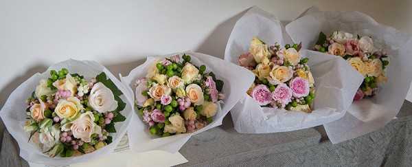 A detail/close-up shot of four bridesmaid or wedding bouquets laid flat on a grey linen surface against a white wall. Each bouquet is wrapped in white tissue paper and contains a soft, pastel arrangement including peach and blush garden roses, dusty pink spray roses, white freesia or similar small blooms, pink waxflower, and clusters of green hypericum berries. The overall floral palette is a mix of peach, blush pink, soft pink, and green, giving a delicate, romantic feel consistent with a classic or garden-style wedding aesthetic.