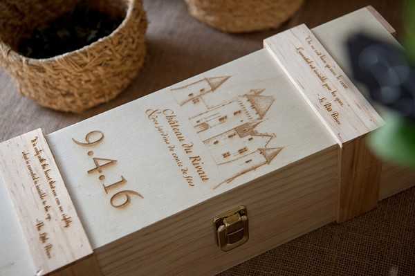 Close-up detail shot of a custom engraved wooden wine box personalized for a wedding. The light pine wood box features laser-engraved text reading '9.4.16' in large numerals alongside an illustration of a château building and the text 'Château de Riven' with additional script writing. The box has a brass latch closure and engraved text visible on the side panel as well, suggesting handwritten letters or vows are stored inside. This appears to be a wine box ceremony keepsake, a popular French wedding tradition where the couple seals wine and letters inside to open on a future anniversary. Woven baskets and blurred greenery are visible in the background on what appears to be a burlap-covered surface.
