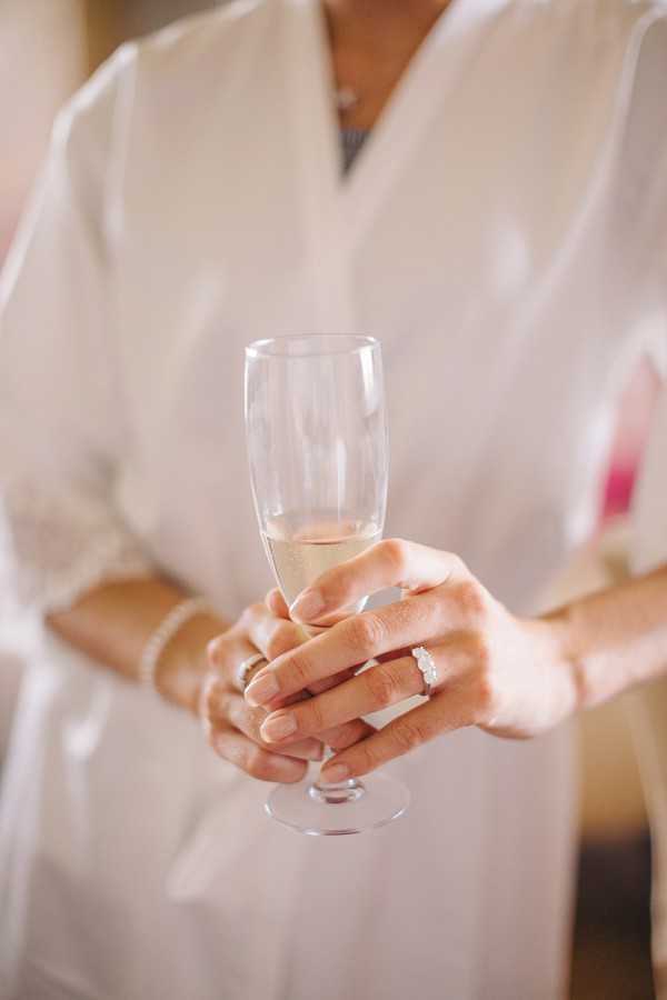 A close-up detail shot taken during bridal preparations, showing a woman's hands holding a champagne flute with a small amount of sparkling wine. She is wearing a white silk or satin robe, a diamond engagement ring on her left hand, and a delicate bracelet on her wrist. A hint of pink — likely a flower or bridesmaid's dress — is visible in the soft-focus background. The overall styling is clean and classic, with a warm, softly lit indoor setting.