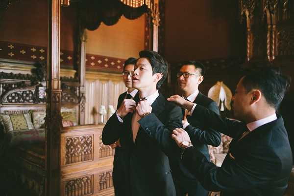 A groom getting ready scene in which two groomsmen help adjust the groom's black bow tie and dark navy suit jacket while a third groomsman stands in the background observing. The setting is an ornately decorated indoor room featuring dark carved wooden furniture, a large mirror with an elaborate wooden frame, and rich jewel-toned upholstery, suggesting a historic chateau or palace interior. All four men are wearing dark navy or black suits with white dress shirts and rose gold watches visible on their wrists. The shot is a medium close-up portrait capturing the candid, preparatory moment from a slightly wide angle.