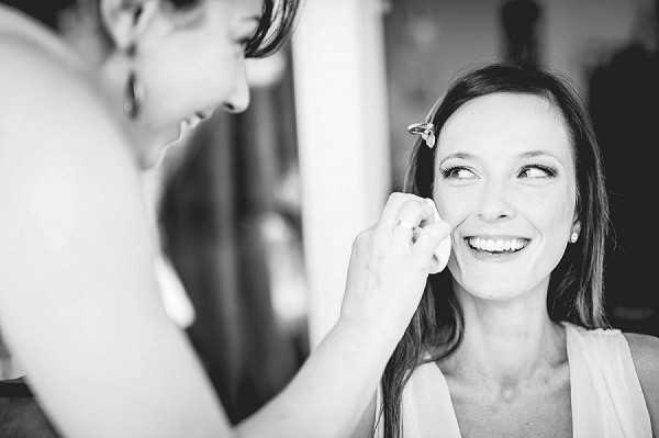A black-and-white getting-ready shot of a bride smiling broadly as a makeup artist applies a finishing touch to her face. The bride wears a small floral or decorative hair clip, stud earrings, and a delicate necklace, with long hair loosely styled. The makeup artist, shown from behind and slightly out of focus, wears a sleeveless top. The image is a close-up portrait with soft contrast and good tonal range, captured in an indoor setting with natural light visible in the background.
