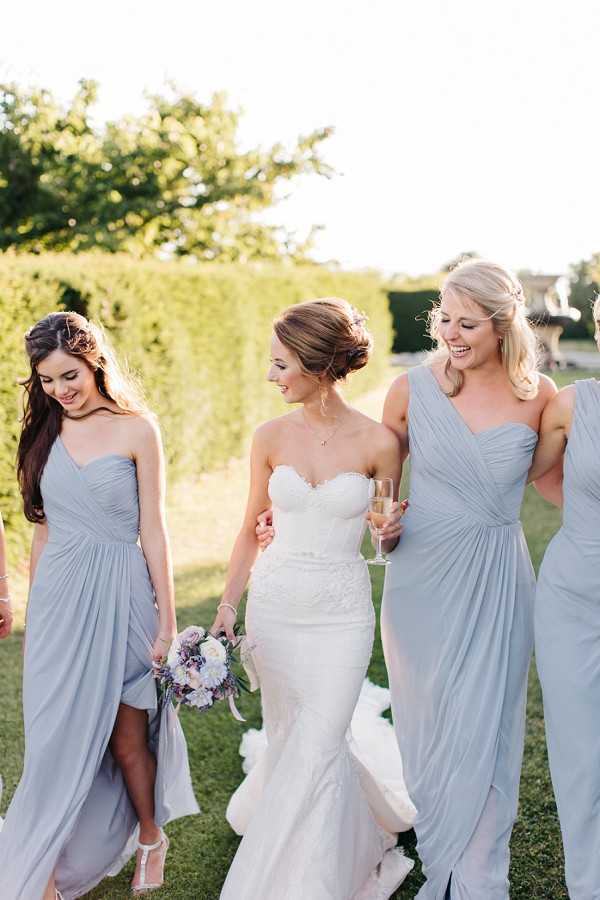 A bridal party portrait taken outdoors in a formal garden setting, showing the bride walking alongside at least three bridesmaids. The bride wears a strapless lace sweetheart-neckline fitted gown and carries a bouquet of white peonies, lavender blooms, and trailing ribbons. The bridesmaids wear floor-length pale silver-blue chiffon gowns in varying neckline styles, including one-shoulder and sweetheart cuts. One bridesmaid holds a champagne flute. The group is laughing and walking casually toward the camera in warm late-afternoon golden light, with a neatly trimmed tall hedge visible in the background. The shot is a medium portrait with a shallow depth of field, keeping the focus on the faces and upper bodies of the women.