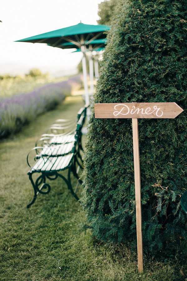 Detail shot of an outdoor wedding directional sign reading 'Dîner' in white hand-lettered calligraphy on a wooden arrow-shaped board mounted on a stake, positioned beside a large trimmed evergreen hedge. In the background, white-slatted garden benches with dark wrought-iron frames are visible alongside teal patio umbrellas, with a row of lavender plants visible in the soft-focus distance. The setting appears to be a French countryside property with a Provençal feel, likely during a summer wedding. The rustic wooden signage style is consistent with a classic French country wedding aesthetic.