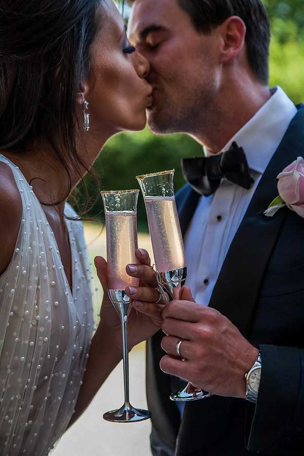 A close-up portrait of a bride and groom sharing a kiss while each holding a champagne flute with gold-rimmed edges containing pink champagne. The bride wears a white gown with pearl embellishments and drop crystal earrings, while the groom is dressed in a navy tuxedo with a black bow tie and a light pink rose boutonniere. Both wear wedding bands, and the groom also wears a silver watch. The shot is tightly framed from roughly the shoulders up, with the champagne flutes prominent in the foreground, set against a softly blurred outdoor background.