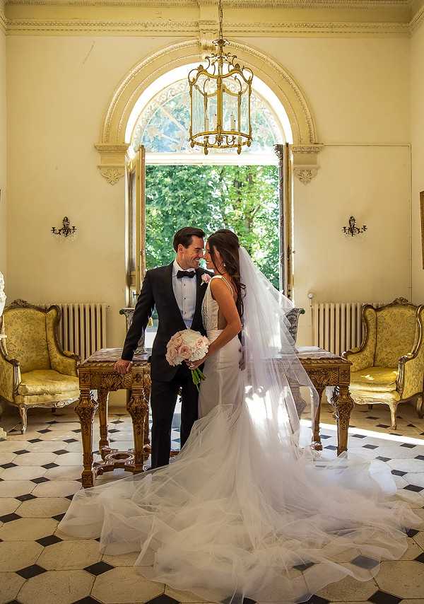 A couple portrait taken indoors in a French chateau or manor house, with the bride and groom standing close together and smiling at each other beneath a large arched window that frames greenery outside. The groom wears a classic black tuxedo with a white dress shirt and black bow tie, while the bride wears a fitted ivory satin gown with a long cathedral-length veil that fans out across the black-and-white hexagonal tile floor. She holds a bouquet of blush and ivory flowers, likely peonies and roses. The room features ornate gold-framed Louis XVI-style armchairs upholstered in yellow-green fabric, a gilded console table, wall sconces, and a gold lantern-style chandelier hanging from an elaborately molded ceiling. The overall styling is classic and formal. Medium full-length portrait shot. Potential venue feature image.