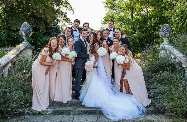 A bridal party group portrait taken on an outdoor stone staircase with ornamental pine cone-topped balustrade finials, likely at a French chateau or formal garden. The bride wears a fitted white gown with a full tulle skirt and long veil, holding a large bouquet of white hydrangeas and pale pink flowers, while the groom wears a navy suit with a black bow tie. Approximately six bridesmaids wear floor-length blush pink multiway dresses and carry round bouquets of white hydrangeas, and roughly six groomsmen wear dark suits with black bow ties. The group of around fourteen people is arranged across several stair steps in a wide group portrait shot, with lavender plantings and manicured greenery visible in the background.