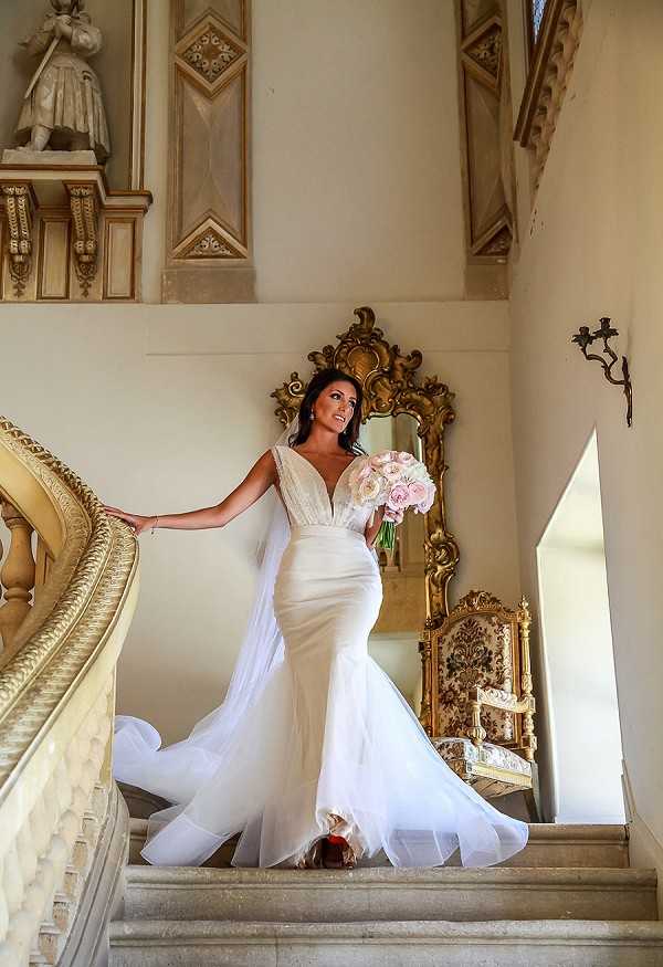 A bridal portrait taken indoors on the grand staircase of a chateau or historic manor, featuring ornate gold-painted banisters, decorative moldings, and a large gilt-framed mirror on the landing. The bride stands alone on the stairs, wearing a fitted white mermaid-style gown with a deep V-neckline, lace bodice details, and a flowing tulle overskirt that trails down the steps. She holds a rounded bouquet of blush and ivory roses, and her dark hair is styled up. Behind her, a gold baroque-style armchair and a wall-mounted candle sconce are visible, reinforcing the classic French chateau interior aesthetic. The shot is a full-length portrait taken from a slightly low angle, capturing the full gown and architectural details of the space. Potential venue feature image.