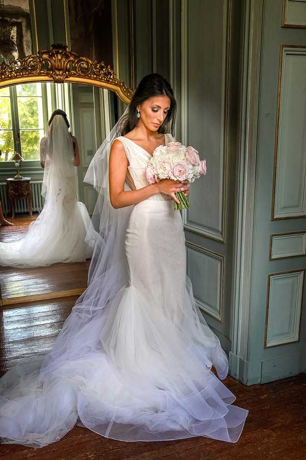 A bridal portrait taken indoors in what appears to be a chateau or historic French venue, featuring ornate sage green wood-paneled walls with gold trim detailing and a large gold-framed mirror. The bride stands in a fitted mermaid-style white tulle gown with a V-neckline, layered tulle skirt hem, and a long cathedral-length veil, looking down at her bouquet of blush pink and ivory garden roses. Her reflection is visible in the mirror behind her, revealing the full train of the dress. The bouquet is a tight round arrangement in soft blush and cream tones. The room has dark hardwood flooring and classic French interior architectural details. Full-length portrait composition with a classic, formal styling theme.