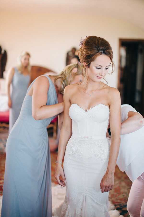 A getting-ready scene indoors, likely in a bedroom or bridal suite, where two bridesmaids in floor-length dusty blue-grey dresses are helping the bride into her gown. The bride is wearing a strapless, sweetheart-neckline white lace fitted dress with a corset bodice and lace appliqué detailing on the skirt, paired with a delicate pendant necklace and a small floral hairpiece with lavender-toned flowers in her upswept brunette hair. A third bridesmaid in the same dusty blue-grey dress is visible in the background, also assisting. The room features warm, soft natural light and a patterned rug on the floor. Medium close-up portrait shot with the bride as the sharp focal point and the background slightly soft-focused.