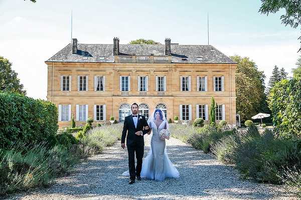 A bride and groom walk arm-in-arm along a gravel path leading away from a large French chateau, facing the camera in a wide portrait shot. The groom wears a black tuxedo with a white dress shirt and black bow tie, while the bride wears a fitted ivory mermaid-style gown with a long veil and holds a white bouquet. The formal chateau is constructed in golden limestone with symmetrical white-shuttered windows across two stories, and the path is flanked by lavender borders and neatly trimmed topiary. Potential venue feature image.