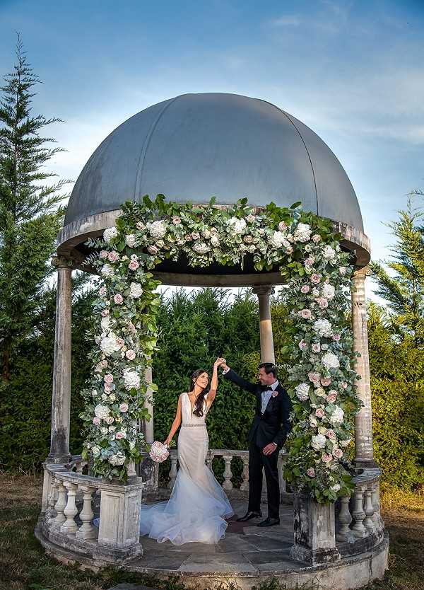 A bride and groom pose together outdoors beneath a stone gazebo with a large dark metal dome roof, photographed in a wide portrait shot. The groom, dressed in a black tuxedo with bow tie, raises the bride's hand as she spins lightly; she wears a fitted white gown with a deep V-neckline, a thin embellished waist detail, and a flowing skirt, and holds a small round bouquet of blush and ivory blooms. The gazebo columns are wrapped in lush floral installations featuring white hydrangeas, blush roses, and abundant green eucalyptus and mixed foliage, continuing across the top rim of the structure. The overall decor palette is white, blush pink, and green, with a classic French formal garden aesthetic. Potential venue feature image.