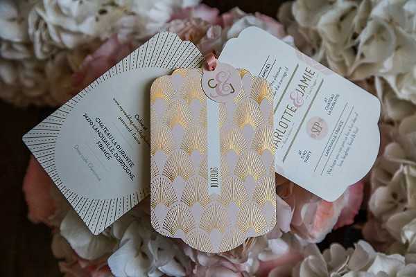 Close-up flat lay of a wedding stationery suite arranged on a bed of blush pink and white hydrangeas. The suite includes three pieces: a main invitation card featuring a blush pink and gold foil Art Deco fan/shell pattern with the date 10.19.16, a details card with a sunburst geometric design in white and gold, and an RSVP card, all printed with the names Charlotte and Jamie. The stationery uses a blush pink, white, and rose gold color palette with Art Deco styling, and the pieces are connected with a small rose gold monogram tag. The venue address visible on the details card references Château La Durantie, Dordogne, France.