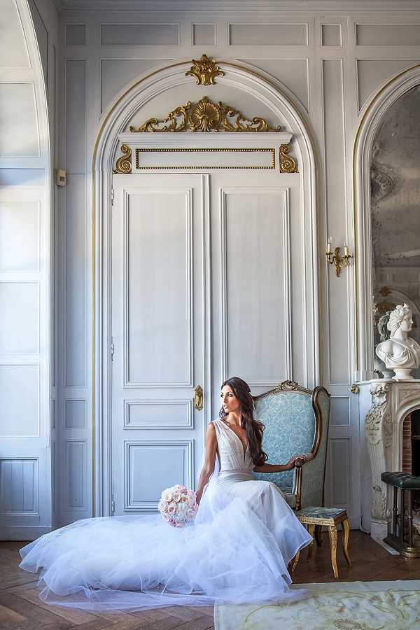A bridal portrait of a single bride seated on the floor beside a gold-framed chair upholstered in pale blue damask fabric, set inside a classically decorated French chateau interior. The bride wears a white tulle ball gown with a deep V-neckline and a voluminous skirt spread across the parquet floor, and holds a rounded bouquet of blush pink peonies or garden roses. The room features white painted boiserie wall paneling with gilded carved ornamental details above a tall arched double door, a gold candle sconce on the wall, and a carved white marble fireplace mantel topped with a classical white bust sculpture. The composition is a full-length portrait shot, with the bride positioned slightly off-center against the architectural backdrop. Potential venue feature image.