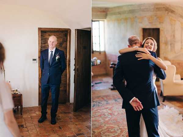 A diptych of two indoor getting-ready or first-look moments set inside what appears to be a historic chateau or villa with terracotta tile floors, aged frescoed walls, and rustic wooden doors. On the left, the groom stands smiling in a navy suit with a small white and greenery boutonnière, hands behind his back, reacting as the bride approaches from the left edge of the frame — wide portrait shot. On the right, the bride in a white gown embraces a man in a navy suit — likely a family member — inside the same interior space furnished with a patterned area rug and cream upholstered chair; the bride is smiling broadly — medium portrait shot. Both images have a light, airy tone with natural window light.