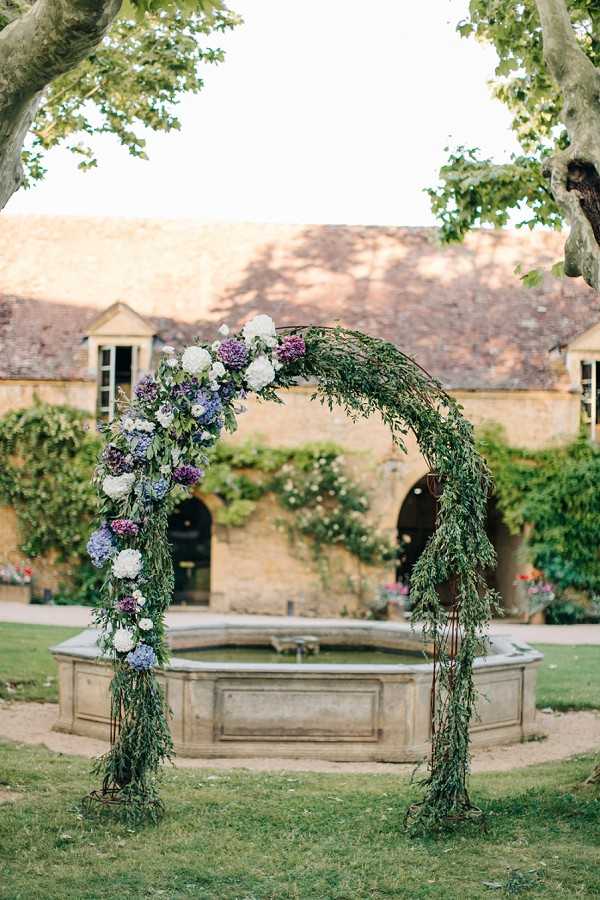 A floral ceremony arch is positioned outdoors in a courtyard in front of a stone fountain, with a ivy-covered French stone building visible in the background. The circular arch is constructed from trailing greenery and adorned primarily on the left side with clusters of blue and purple hydrangeas, white peonies, and small purple blooms. No people are present; this is a decor detail shot capturing the ceremony setup. The overall palette is purple, blue, and white against lush green foliage, set within what appears to be a classic French country estate courtyard. Potential venue feature image.