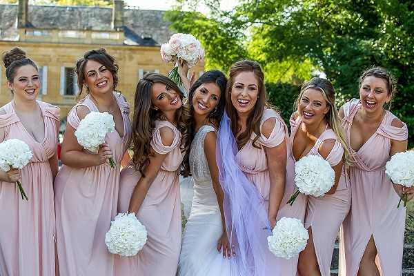 A bridal party portrait taken outdoors featuring the bride and six bridesmaids posing closely together and laughing. The bride wears a white fitted gown with beaded detailing and a long white veil, while all six bridesmaids wear matching blush pink floor-length multiway wrap dresses with a cold-shoulder style. The bridesmaids each carry round bouquets of white hydrangeas, and the bride raises her bouquet of white peonies in the air. The shot is a close-up group portrait taken at mid-distance, with a stone manor building and green trees visible in the background.