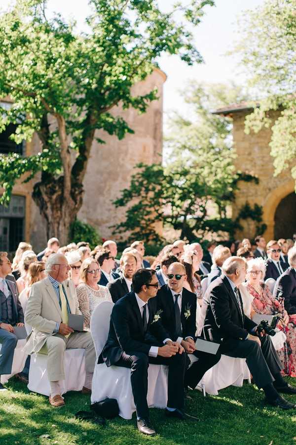 An outdoor wedding ceremony in progress, with approximately 40-50 guests seated in white chair-covered rows on a lawn. In the foreground, two men in dark navy suits with small white floral boutonnieres are seated in the front row and appear to be conversing quietly — these may be groomsmen or the grooms. Guests are dressed in a mix of formal attire including dark suits and summer dresses in floral and light tones. The setting is a courtyard or garden of a historic stone building with arched architectural details visible in the background. The ceremony area is shaded by large mature trees, with bright midday sunlight creating dappled light across the scene. Wide shot taken from a slightly elevated angle looking back toward the seated guests.