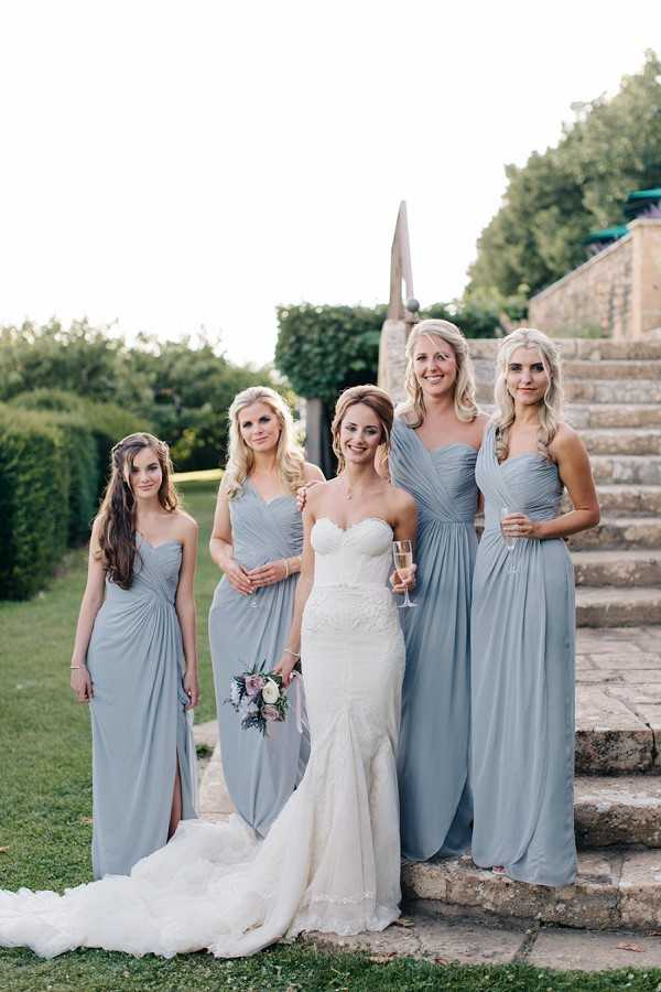 A bridal party portrait taken outdoors on stone steps, featuring the bride and four bridesmaids posing together. The bride wears a fitted ivory strapless lace gown with a train and holds a small bouquet of blush, mauve, and white blooms with trailing ribbon. The four bridesmaids wear floor-length dusty blue-grey draped chiffon dresses in a sweetheart neckline style, with two of them holding champagne flutes. The setting appears to be the terraced grounds of a chateau or manor property, with stone balustrade steps visible in the background. The overall styling is classic and polished, with a muted blue-grey and ivory color palette.