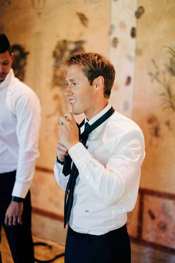 A getting-ready moment captured indoors, showing a groom in a white dress shirt and black tie adjusting his tie with both hands, with a slight smile on his face. A second man in a white dress shirt is partially visible on the left edge of the frame. The background features an ornate painted or frescoed wall with botanical motifs in warm ochre and cream tones, suggesting a historic chateau or villa interior. The shot is a close-up portrait with warm ambient lighting.