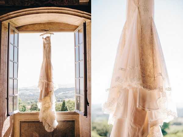A getting-ready detail shot presented as a diptych, showing a wedding dress hanging from an ornate arched window inside what appears to be a chateau or historic venue. The left panel shows the full-length gown suspended on a hanger against the open double French doors, with a countryside landscape visible in the background. The right panel is a close-up detail of the dress fabric, revealing a blush/champagne-toned gown with tiered layers, delicate lace trim, and gold embroidered floral detailing along the hem and bodice. The dress has a fitted silhouette with a flowing layered skirt. The window frame features carved stone or painted wood detailing consistent with a historic French property.