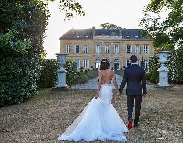 A couple portrait shot from behind showing the bride and groom walking hand-in-hand toward a large French chateau along a gravel path at golden hour. The bride wears a fitted white mermaid-style gown with a low open back, a cathedral-length train, and a decorative hair piece, while the groom wears a dark navy suit with red-soled shoes visible mid-stride. The chateau facade is built in honey-colored stone with symmetrical shuttered windows, flanked by two ornamental stone urns on pedestals and formally trimmed hedges. Wide shot composition with the chateau as the central focal point. Potential venue feature image.