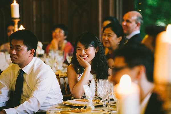 Wedding reception dinner scene shot indoors, likely during speeches or toasts, with guests seated at a long dining table. A woman in the foreground wearing a patterned strapless dress is smiling broadly with her hand resting against her cheek, while a man beside her in a white dress shirt and dark tie looks toward the same focal point. Several other guests are visible in the background, including an older woman in a red top and a man in a dark suit. The table is set with gold charger plates, glassware, and candles, and tall pillar candles on candlesticks are visible in the background, casting warm amber light throughout the darkened interior space. The setting appears to be a rustic or historic hall with dark wood paneling. Medium-wide candid shot capturing a genuine moment of enjoyment among guests.