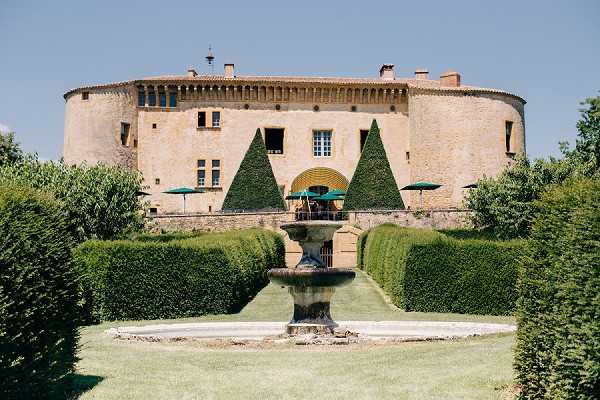 Wide shot of a French chateau exterior with formal garden grounds in the foreground. The building is a round-turreted stone chateau with warm beige masonry, visible windows, and chimney stacks along the roofline. The formal garden features precisely clipped conical topiary hedges, a central stone fountain with a tiered basin, and symmetrically trimmed tall hedgerows framing a central lawn pathway. A terrace area between the garden and the chateau entrance shows green market umbrellas, suggesting a cocktail or reception setup. No people are visible in the frame. Potential venue feature image.