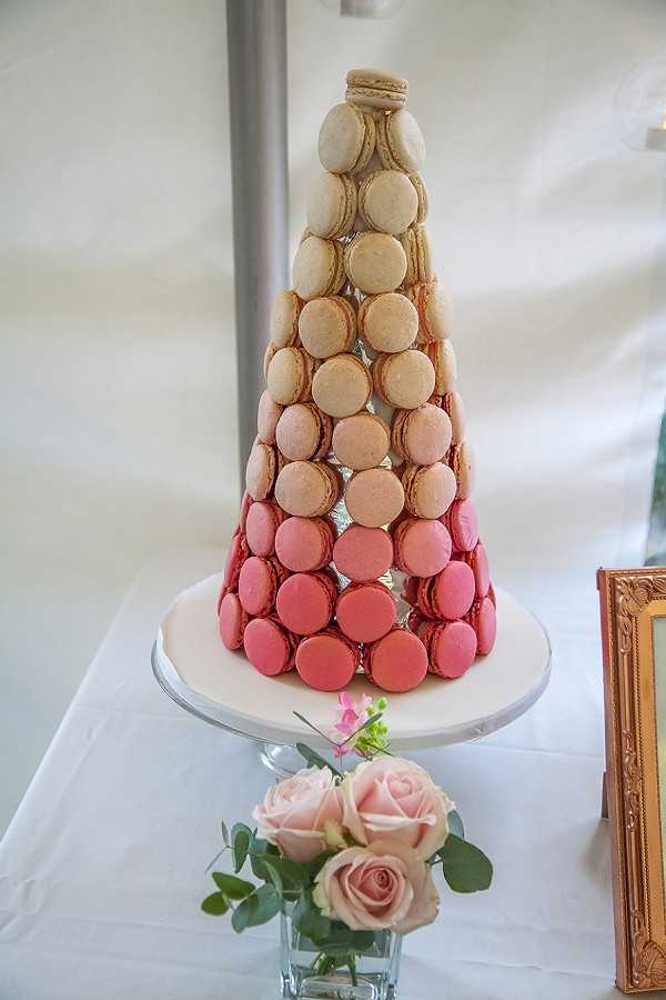 Close-up detail shot of a French macaron tower displayed at a wedding reception dessert table. The tower is arranged in a cone shape on a white pedestal cake stand, featuring an ombré color gradient that transitions from deep raspberry and coral pink at the base through blush and pale peach tones to cream and vanilla at the top. In front of the stand sits a small glass vase with blush pink roses and eucalyptus sprigs. A gold-framed item is partially visible to the right. The table is covered with a white linen, and the background suggests a tented or draped white reception space.