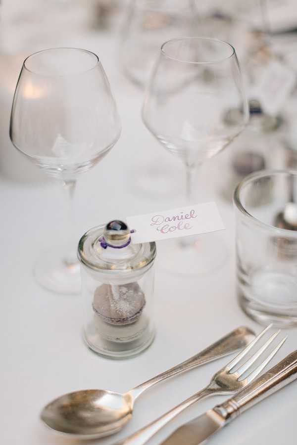Close-up detail shot of a wedding reception place setting on a white linen tablecloth. A small glass jar containing a lavender-grey macaron serves as a guest favor, topped with a small decorative stopper and a white place card with the name 'Daniel Cole' written in pink script lettering. The setting includes silver cutlery (spoon, knife, and fork), two clear wine glasses, and a tumbler glass visible to the right. The overall decor palette is white and pale grey with soft purple accents, reflecting a clean, minimal styling approach. Additional place settings are softly blurred in the background.