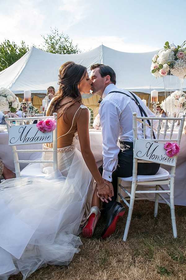 The bride and groom are seated at their sweetheart table kissing during an outdoor reception held beneath a large white stretch tent. Their white chiavari chairs are decorated with calligraphy signs reading 'Madame' and 'Monsieur,' each adorned with a hot pink peony. The bride wears a backless white gown with a beaded belt and tulle skirt, with red-soled heels visible beneath; the groom wears a white dress shirt with black suspenders and black trousers. Tall centerpieces of white and blush flowers — including peonies and hydrangeas — are visible on white-linen tables in the background, along with pillar candles, and a group of guests can be seen seated around the reception tables. The overall decor palette is white, blush, and hot pink with a classic French-inspired style. Medium-distance portrait shot.