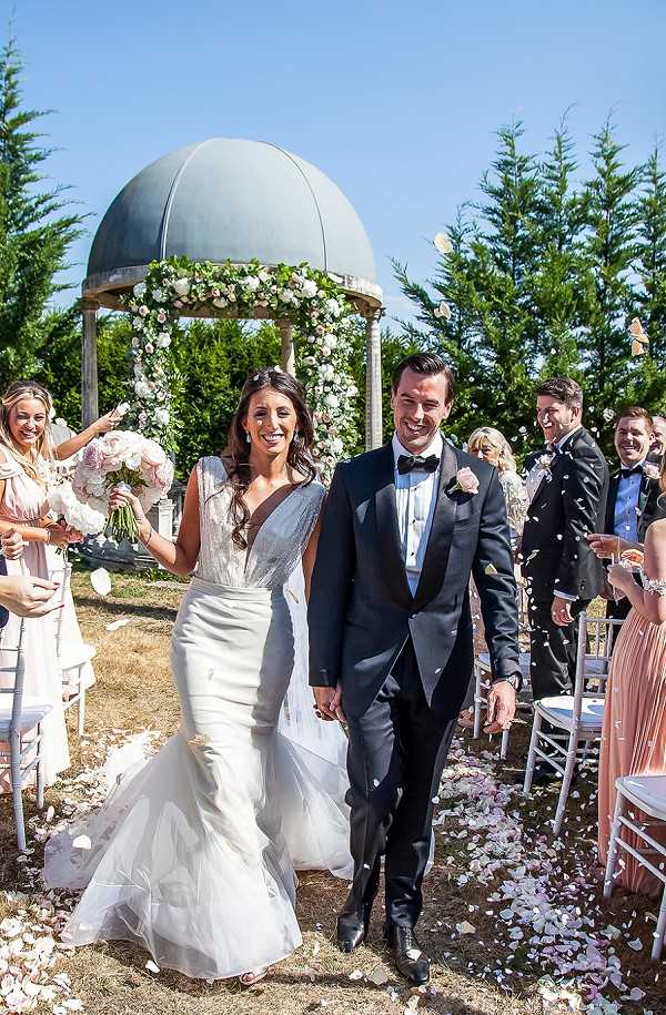 The bride and groom walk back up the aisle following an outdoor ceremony, with guests on either side tossing white and blush pink flower petals over them. The bride wears a fitted mermaid-style white gown with a deep V-neckline and carries a bouquet of blush pink and white roses, while the groom is dressed in a dark navy tuxedo with a black bow tie and a blush pink buttonhole. Behind them stands a classical stone gazebo with a teal domed roof, decorated with a floral arch of white and blush pink roses with greenery. Guests visible in the background and along the aisle include women in blush pink dresses and men in dark suits, seated on white chiavari chairs; flower petals are scattered along the aisle. The overall styling is classic and polished, with a blush and white floral palette. Portrait-style shot taken from a low angle, capturing the full scene from the couple in the foreground to the gazebo behind.