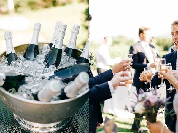 A two-panel detail and candid shot from an outdoor wedding cocktail hour. The left panel shows a close-up of a silver ice bucket filled with multiple chilled champagne bottles on ice, shot with a shallow depth of field. The right panel captures guests in navy suits raising champagne flutes in a toast, with a soft-focus bouquet of lavender and purple flowers visible in the foreground and other guests gathered on a lawn in the background. The overall styling suggests a classic, understated aesthetic with a muted floral palette.