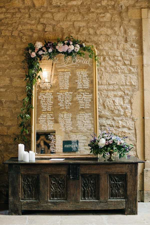 A wedding seating chart display featuring a large gold-framed mirror with white calligraphy script reading 'Please Be Seated' at the top, followed by guest names organized by table. The mirror is decorated along the top and left side with a garland of greenery, blush pink roses, and mauve/dusty purple blooms. The mirror rests on a dark carved antique wooden chest, which also holds three white pillar candles of varying heights on the left and a floral arrangement of white peonies, lavender, purple and mauve blooms with greenery in a brass or gold vessel on the right. The setting is indoors against a rustic exposed limestone or sandstone wall, consistent with a chateau or historic French venue. The overall decor palette is blush, mauve, purple, and gold with a classic-romantic styling. Wide portrait-orientation shot.