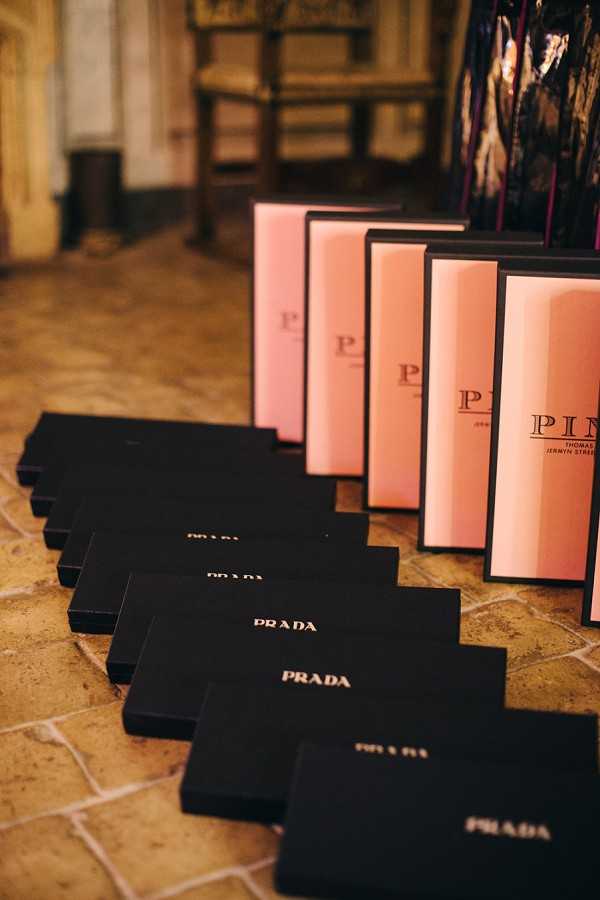 A detail close-up shot of wedding guest gifts arranged in two parallel diagonal rows on a stone tile floor inside what appears to be a historic indoor venue. One row consists of matte black Prada gift boxes with gold foil lettering, and the other row features pink and black framed Pink's of Jermyn Street boxes. Approximately eight to ten of each box type are visible, receding into the background. The overall gift presentation palette is black, gold, and blush pink, suggesting a high-end, designer-focused gifting theme.