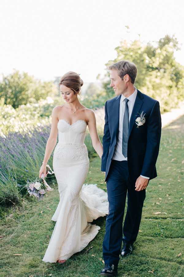 A couple portrait taken outdoors showing the bride and groom walking together and holding hands along a garden path bordered by lavender. The bride wears a fitted ivory strapless lace corset-style gown with a trailing train and carries a loose bouquet of white and pale blush blooms with lavender accents. The groom wears a navy suit with a grey tie and a white and lavender boutonniere. The portrait is shot at a medium distance with warm late-afternoon light and a classic, clean styling approach.
