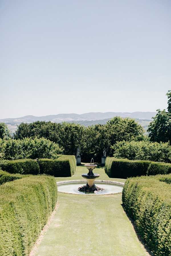A wide shot of formal garden grounds featuring a central stone fountain with a tiered basin set within a circular pool, flanked by two symmetrical rows of precisely clipped tall hedges forming a long allée. Stone pedestals with decorative urns or statues are visible in the background between the hedgerows, and a rolling hillside landscape extends into the distance. No people are present in the frame. The image is taken in bright midday light with a portrait orientation, emphasizing the formal French garden layout. Potential venue feature image.