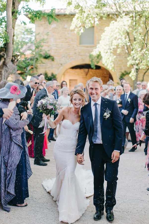 A newly married couple walks through a confetti exit outside a rustic stone building with an arched doorway, likely a chapel or mas in the south of France. The bride wears a fitted strapless lace gown with a short train and carries a loose bouquet of lavender, dusty mauve, and white blooms with trailing ribbons, while the groom wears a navy suit with a light blue tie and a small floral buttonhole. Approximately 30 guests line both sides of the gravel path, cheering and throwing white confetti, with one guest prominently wearing a wide-brim grey hat with a pink flower. The shot is a portrait-orientation mid-range photo with the couple centered in the foreground and the stone venue facade visible in the background.