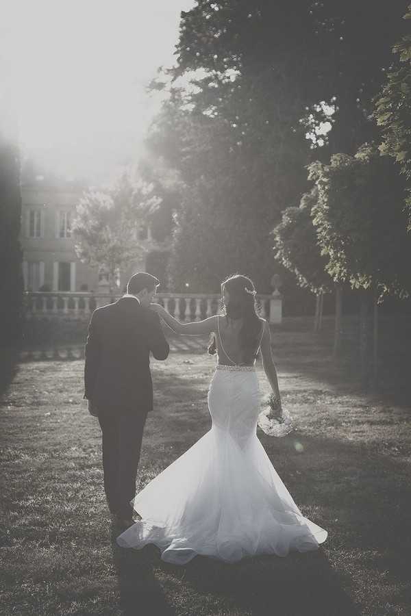 A black-and-white couple portrait shot from behind, showing the bride and groom walking together across a lawn toward a chateau or manor house visible in the background. The groom wears a dark suit and the bride wears a fitted mermaid-style wedding dress with an open back, an embellished waistband, and a flared tulle skirt with a short train; she carries a small bouquet and has her hand resting on the groom's arm. The image is strongly backlit, creating high contrast with bright light flooding the center of the frame and darker tones framing the edges. A stone balustrade is visible near the chateau entrance, lending a classic French estate atmosphere to the setting. Wide portrait shot with a romantic, hazy quality from the strong backlighting.