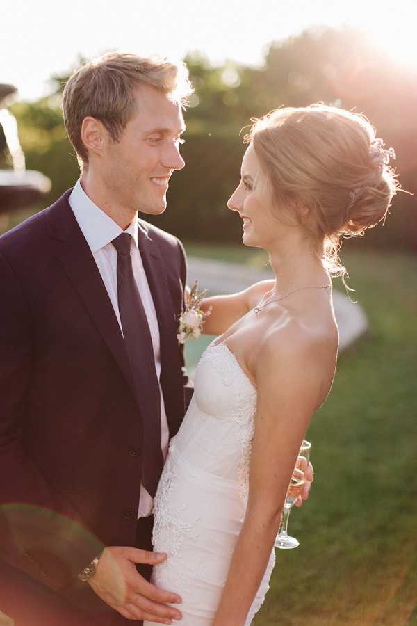 An outdoor couple portrait taken during golden hour, with warm backlit sunlight creating a soft lens flare across the frame. The groom wears a dark navy suit with a dark tie and a small boutonniere featuring blush and white flowers, while the bride wears a strapless ivory lace fitted gown with a delicate necklace and her hair styled in a loose updo. The bride holds a champagne flute and the couple face each other, smiling, in a close semi-profile composition. The styling is classic and understated.