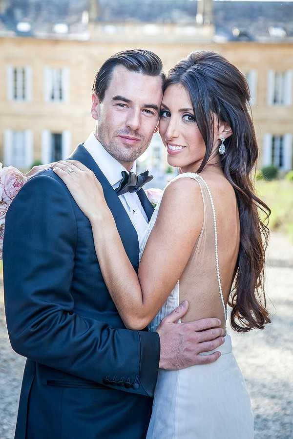 A couples portrait taken outdoors in front of a French chateau, with the stone facade and symmetrical windows of the building visible in the background. The groom wears a navy tuxedo with a black bow tie and white dress shirt, while the bride wears a low-back ivory gown with a pearl-embellished spine detail and teardrop crystal earrings. The bride, with long dark hair styled in a half-up wave, looks back over her shoulder toward the camera while the groom faces forward, their cheeks touching as they embrace. The composition is a close portrait shot with a shallow depth of field that softly blurs the chateau behind them.