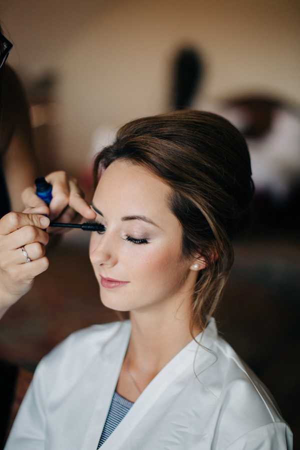 A close-up portrait shot of a bride getting ready, with a makeup artist applying mascara to her lower lashes. The bride has brown hair styled in a soft updo with loose face-framing pieces, and is wearing a white robe over a striped top. Her makeup features neutral eyeshadow, defined lashes, and a soft pink lip. The setting is indoors with a softly blurred background, and the image has a warm, natural light tone. The composition is a tight portrait focused on the bride's face.