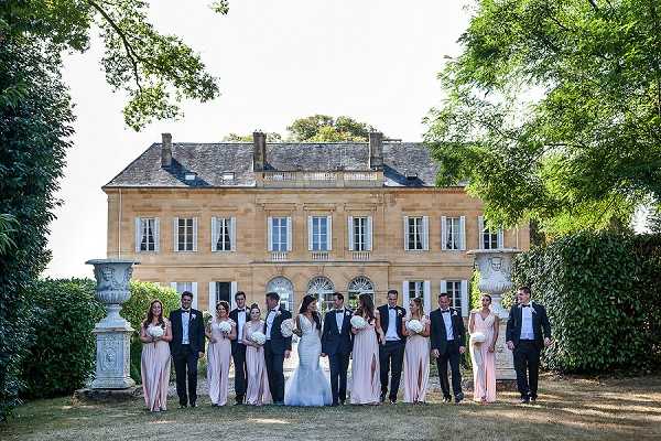 A bridal party group portrait taken outdoors on the grounds of a French chateau, with the golden stone facade of the manor house serving as the backdrop. The bride wears a fitted white strapless gown and carries a white bouquet, while the groom and groomsmen are dressed in dark navy suits with black bow ties and white pocket squares. The bridesmaids wear floor-length blush pink gowns, and the group of approximately twelve people is arranged in a loose line facing the camera in a wide shot. Two large stone decorative urns flank the group symmetrically on the lawn in front of the chateau. Potential venue feature image.