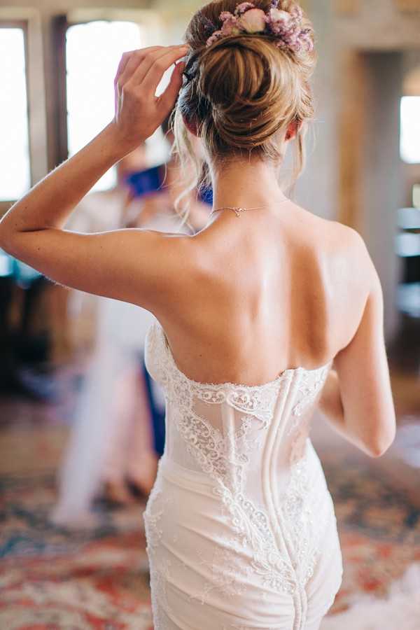 A close-up portrait of a bride shot from behind during the getting-ready phase, taken indoors in a softly lit room. The bride wears a fitted ivory strapless lace wedding dress with an open back, intricate floral lace detailing running down the corset, and a delicate back necklace with a small pendant. Her hair is styled in a loose updo adorned with a floral crown featuring small mauve and blush blooms. She is raising one hand to adjust her hair, and a blurred figure in a blue outfit is visible in the background.
