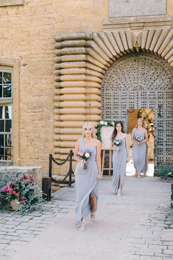 Three bridesmaids walk in procession through a large ornate arched doorway of a historic chateau or manor house, moving toward the camera in an outdoor courtyard setting. All three wear floor-length dusty blue-grey one-shoulder draped dresses with front slit detailing, paired with strappy nude heels. Each bridesmaid carries a small bouquet featuring blush pink and white blooms with dusty purple accents, likely including roses and thistles. The architectural doorway features distinctive rusticated stone detailing with alternating ribbed quoins, and a large decorative floral arch arrangement is visible behind them at the entrance. The image is a medium wide portrait shot with natural daylight, and the overall styling palette is soft grey-blue and blush in a classic, refined aesthetic.